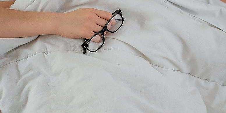 person lying on bed while covering face with pillow and holding eyeglasses