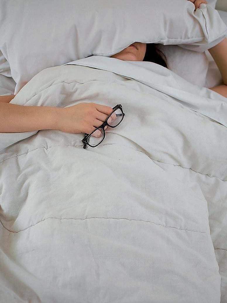 person lying on bed while covering face with pillow and holding eyeglasses